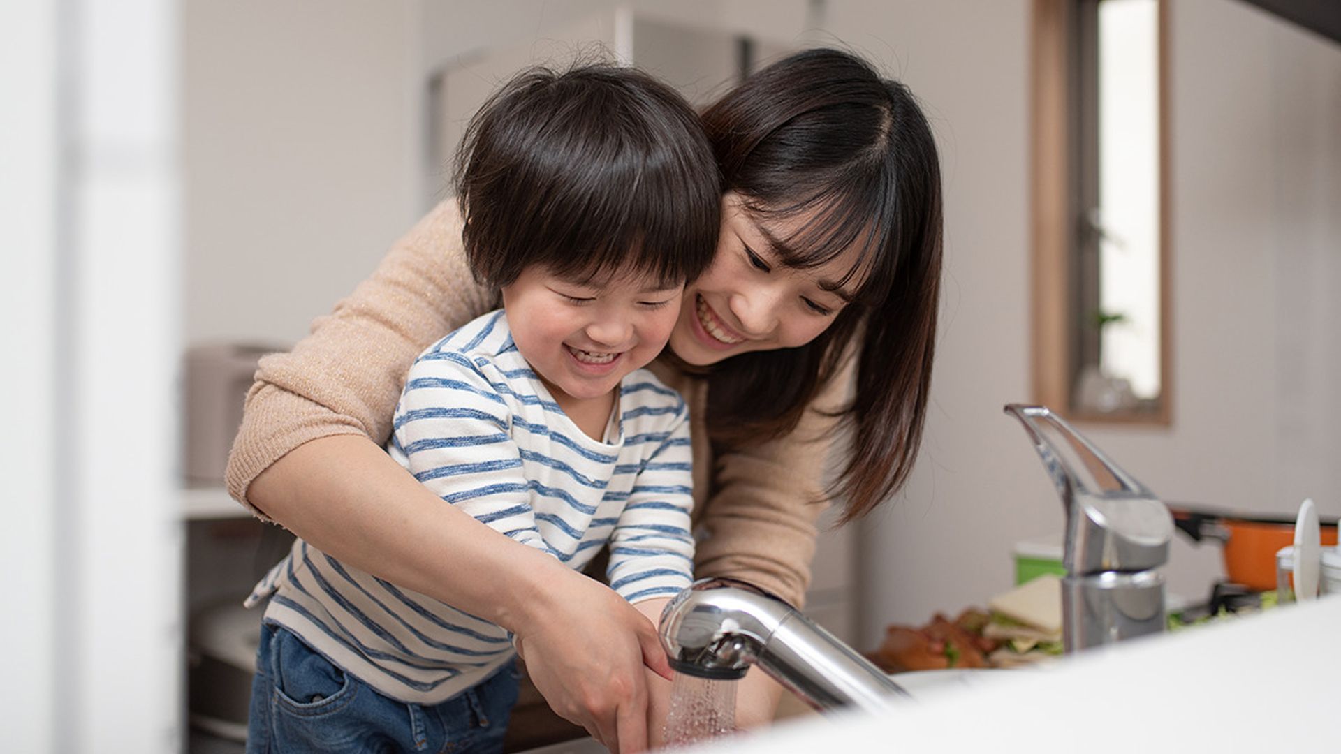 A mom and her young kid are washing their hands in the sink. Both are smiling.
