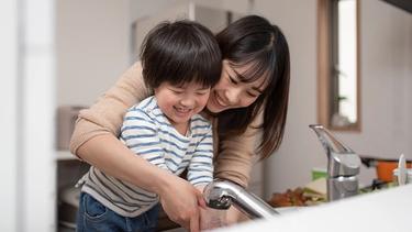 A mom and her young kid are washing their hands in the sink. Both are smiling.