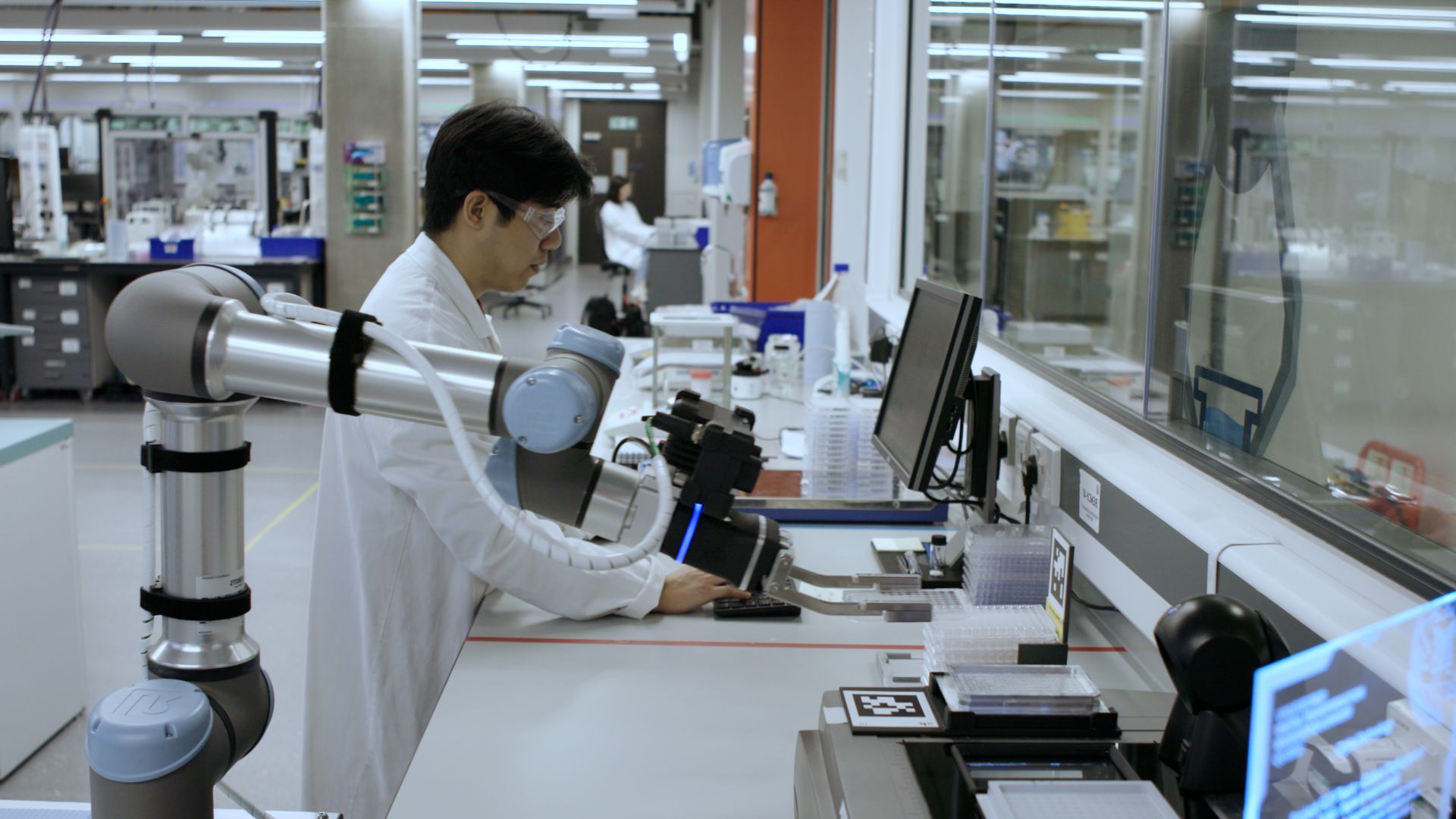 A scientist in a white coat standing in a laboratory, surrounded by various scientific equipment.
