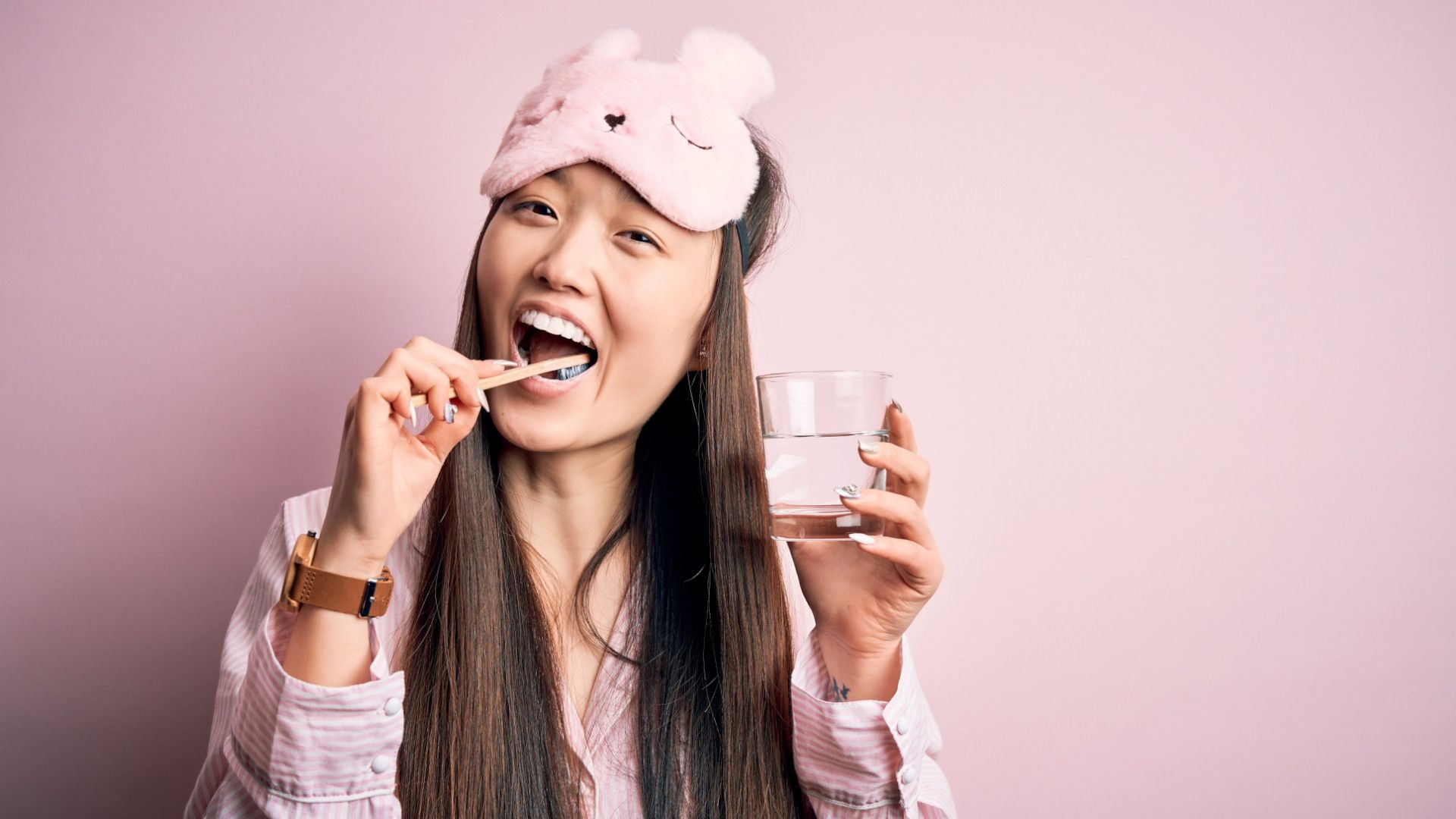 A woman in pajamas and a pink sleep mask is brushing her teeth, smiling, and holding a glass of water against a pink background, conveying a cheerful tone.