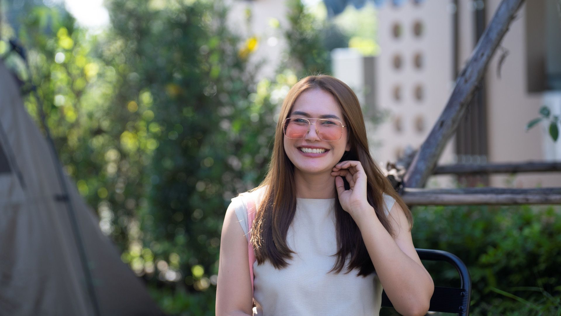 A woman with long brown hair wearing a sleeveless white top and pink sunglasses, sitting outdoors with greenery in the background.