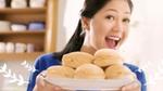 Woman smiling and holding a plate with a stack of round cream-filled buns in a bright kitchen setting.