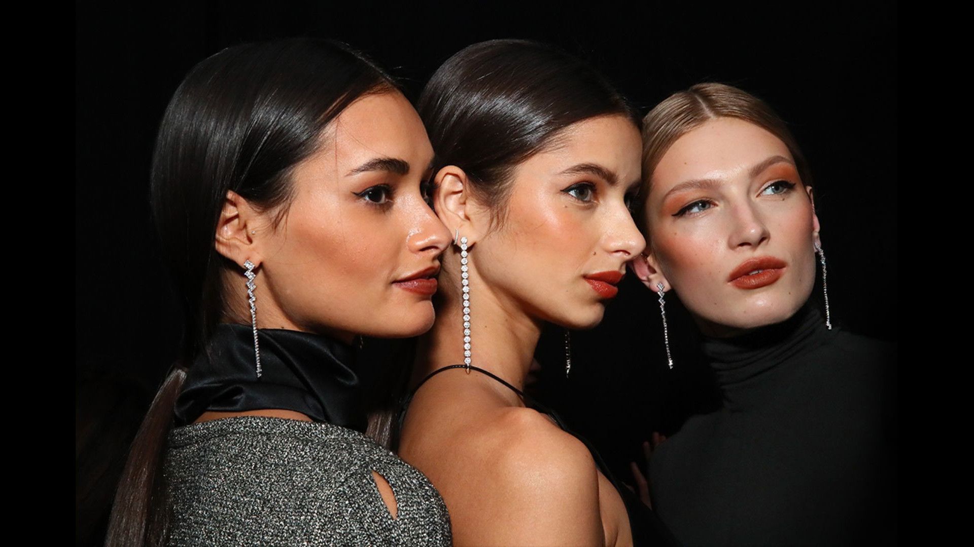 Three women closely posing, wearing elegant black outfits and long dangling earrings, against a dark background.