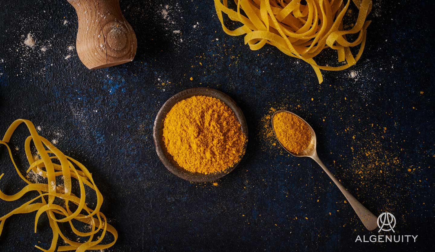 A small bowl of orange microalgae powder on a blue-grey table with two tangles of thick pasta on the side.