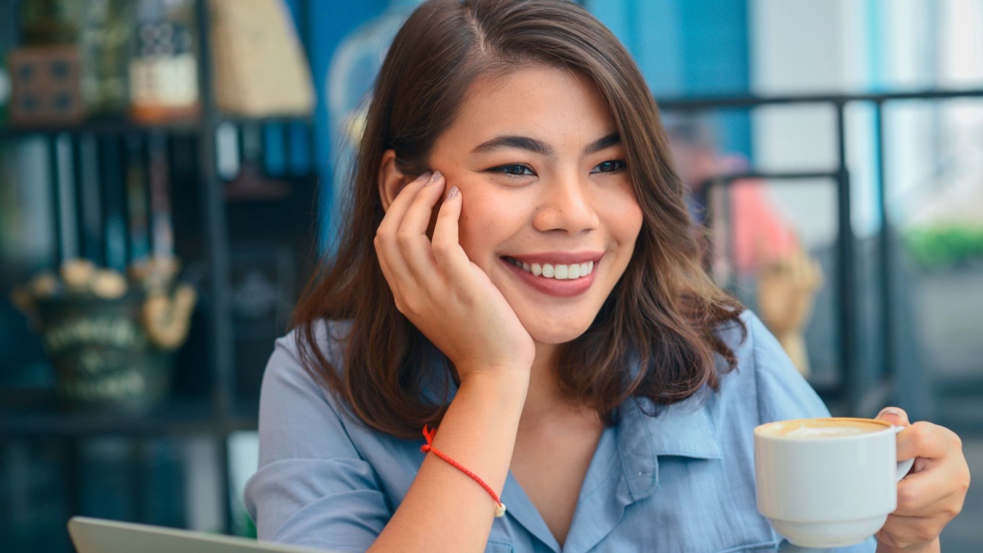 Smiling woman in a blue shirt sits at a cafe, holding a coffee cup. She looks relaxed and content, with a blurred background of shelves.