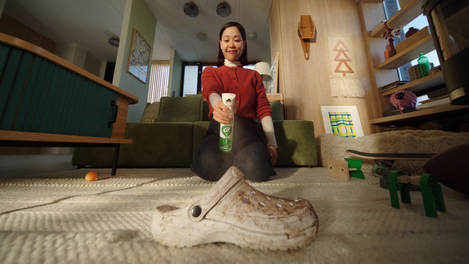 A person is kneeling on a carpeted floor, holding a can of Cif Infinite Clean aimed at a dirty white shoe in the foreground. The room has a cozy decor with a green couch, wooden furniture, and various items scattered around.