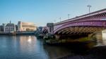 View of Blackfriars Bridge spanning the River Thames with the prominent 100 Victoria Embankment building in the background. The building features a classic architectural style with large windows and a light stone facade, illuminated by bright morning sunlight under a clear blue sky.