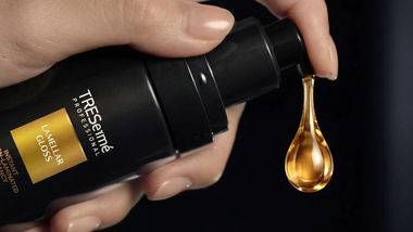 A golden droplet is dispensed by hand from a bottle of TRESemmé Professional Lamellar Gloss Ultra-Gloss Oil.