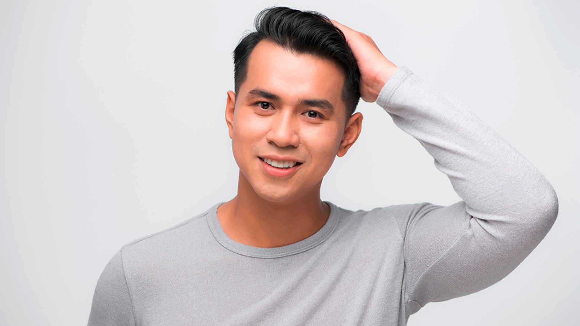 A man in a light gray long-sleeve shirt with one hand on his head against a plain light background.