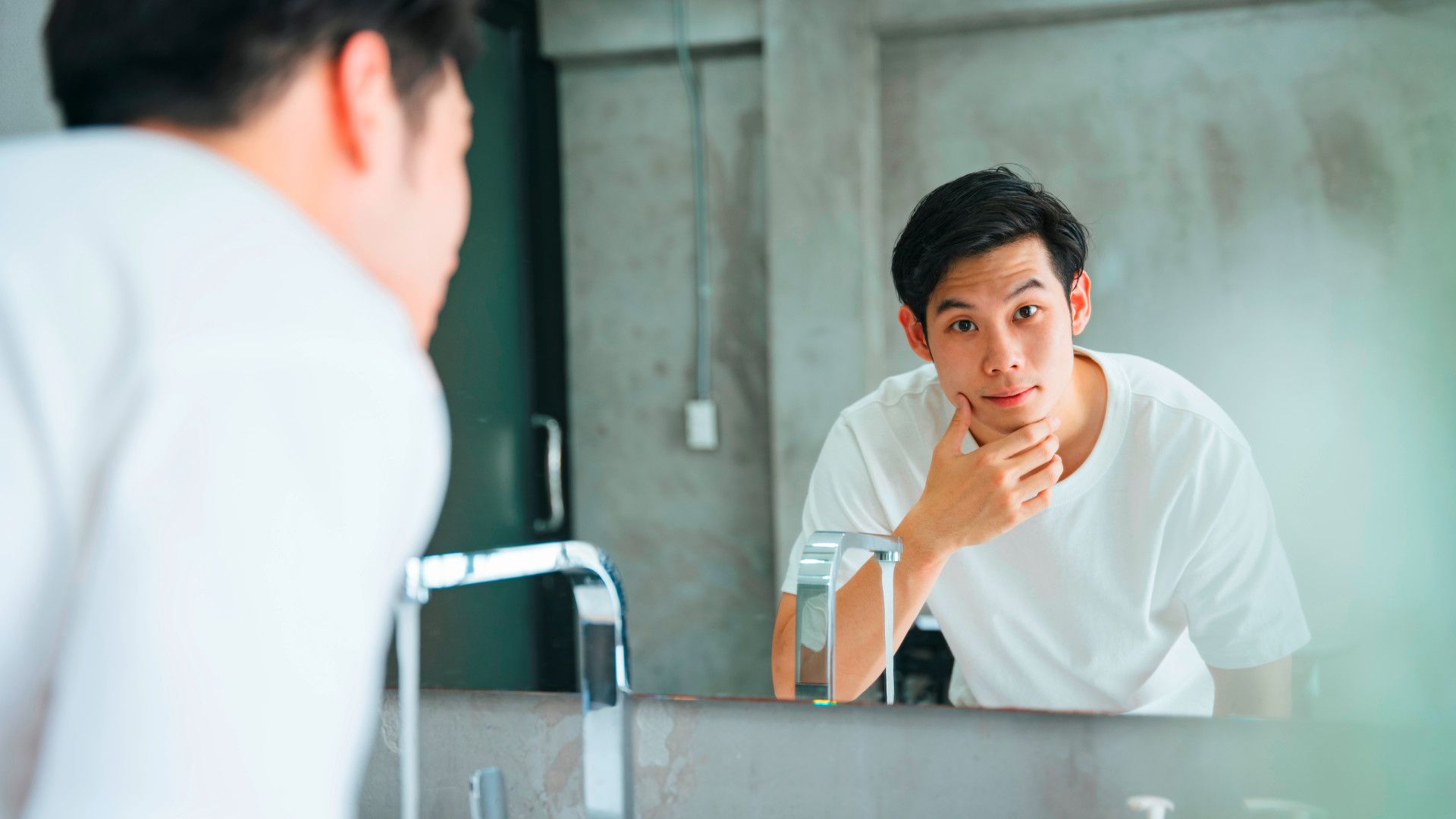 A man in a white t-shirt looking in the mirror.