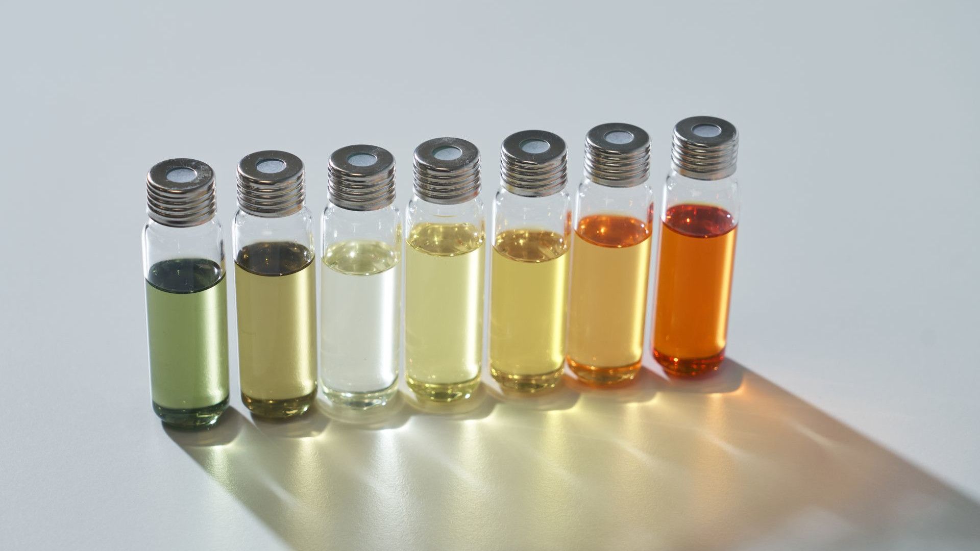 Eight small glass vials containing liquids of varying colors, ranging from green to orange, arranged in a row on a light surface.