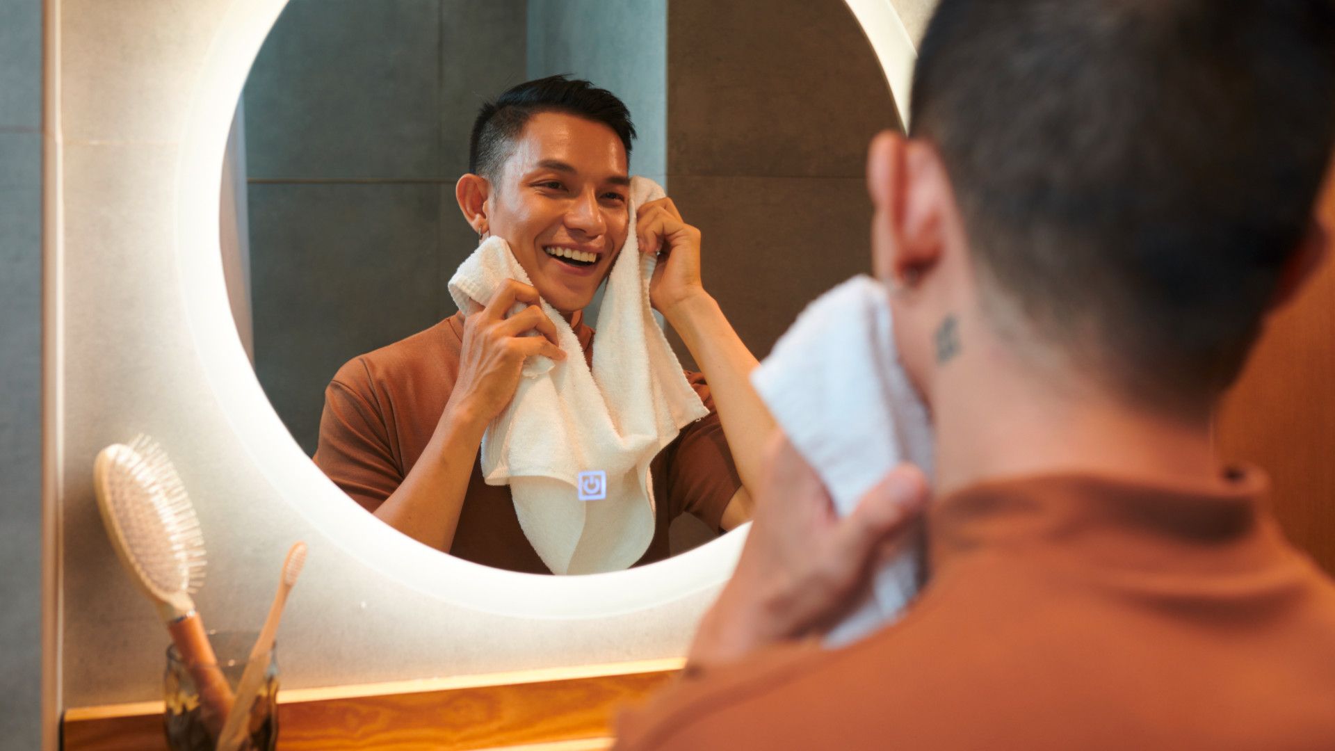 A man smiles while drying his face with a towel in front of a round, illuminated mirror.