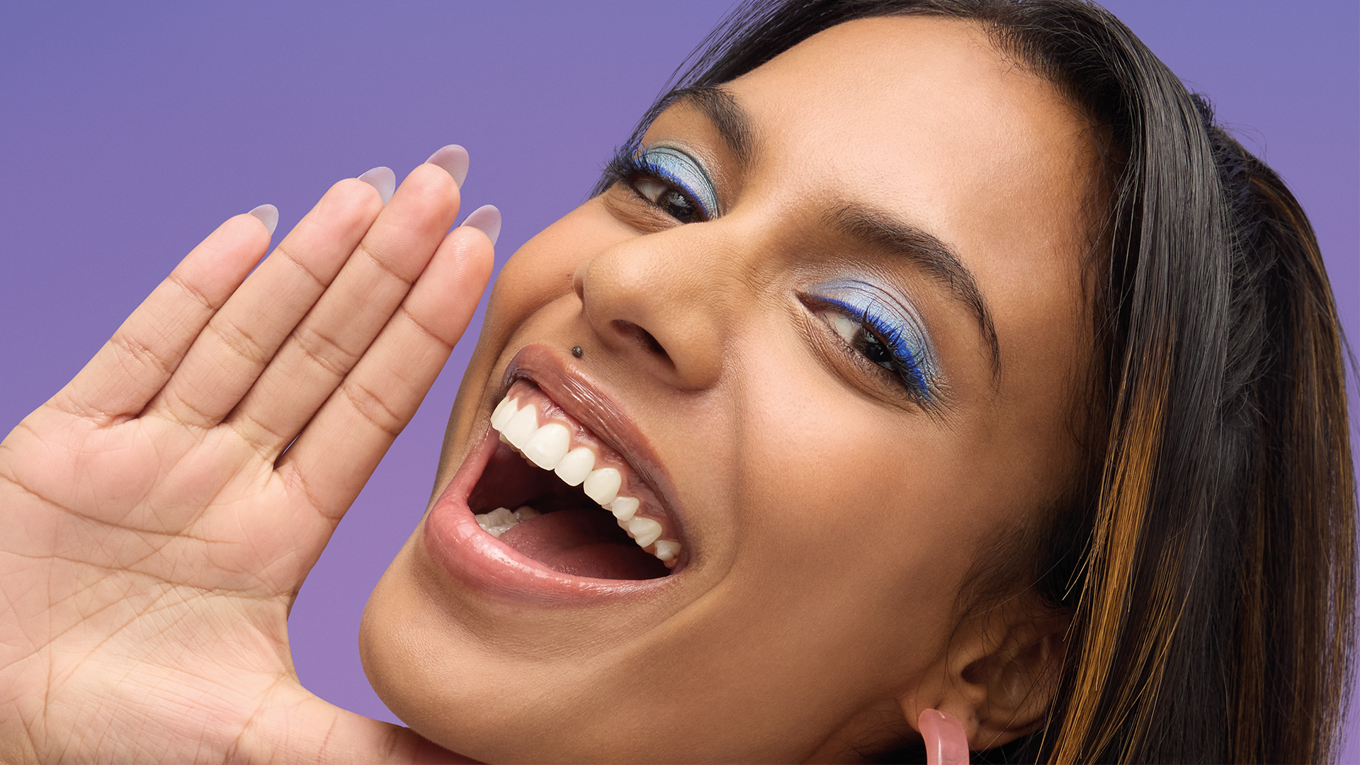 A woman with long dark hair and blue eye makeup smiles brightly against a purple background. She holds one hand up beside her face, showcasing her white teeth and happy expression.