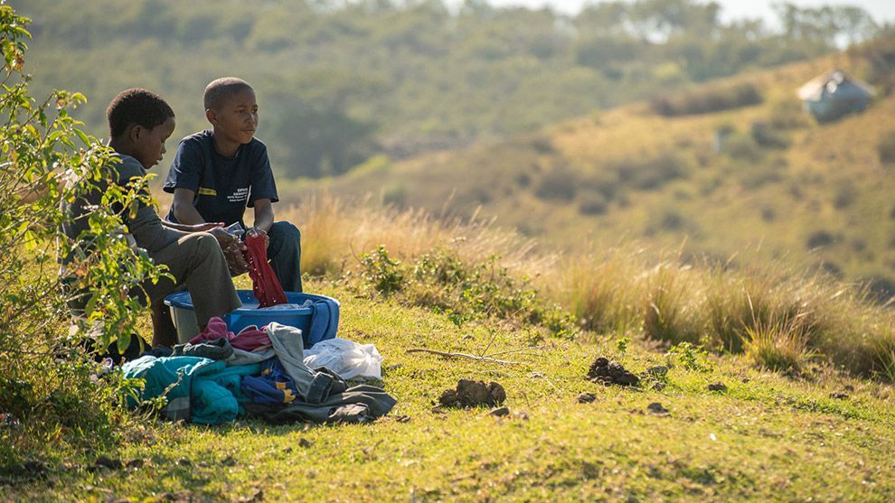 Two boys sit in a field washing clothes over a basin with soap and water