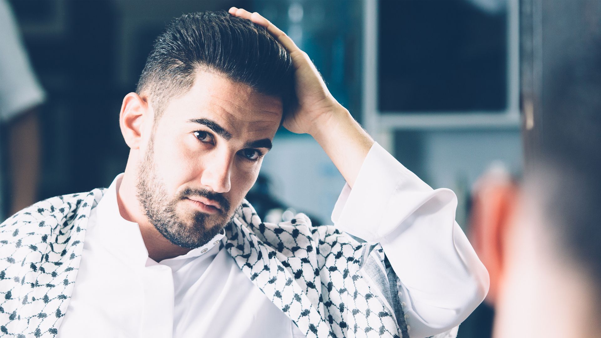 A man in the bathroom has a towel on one shoulder, about to wipe his face while smiling.