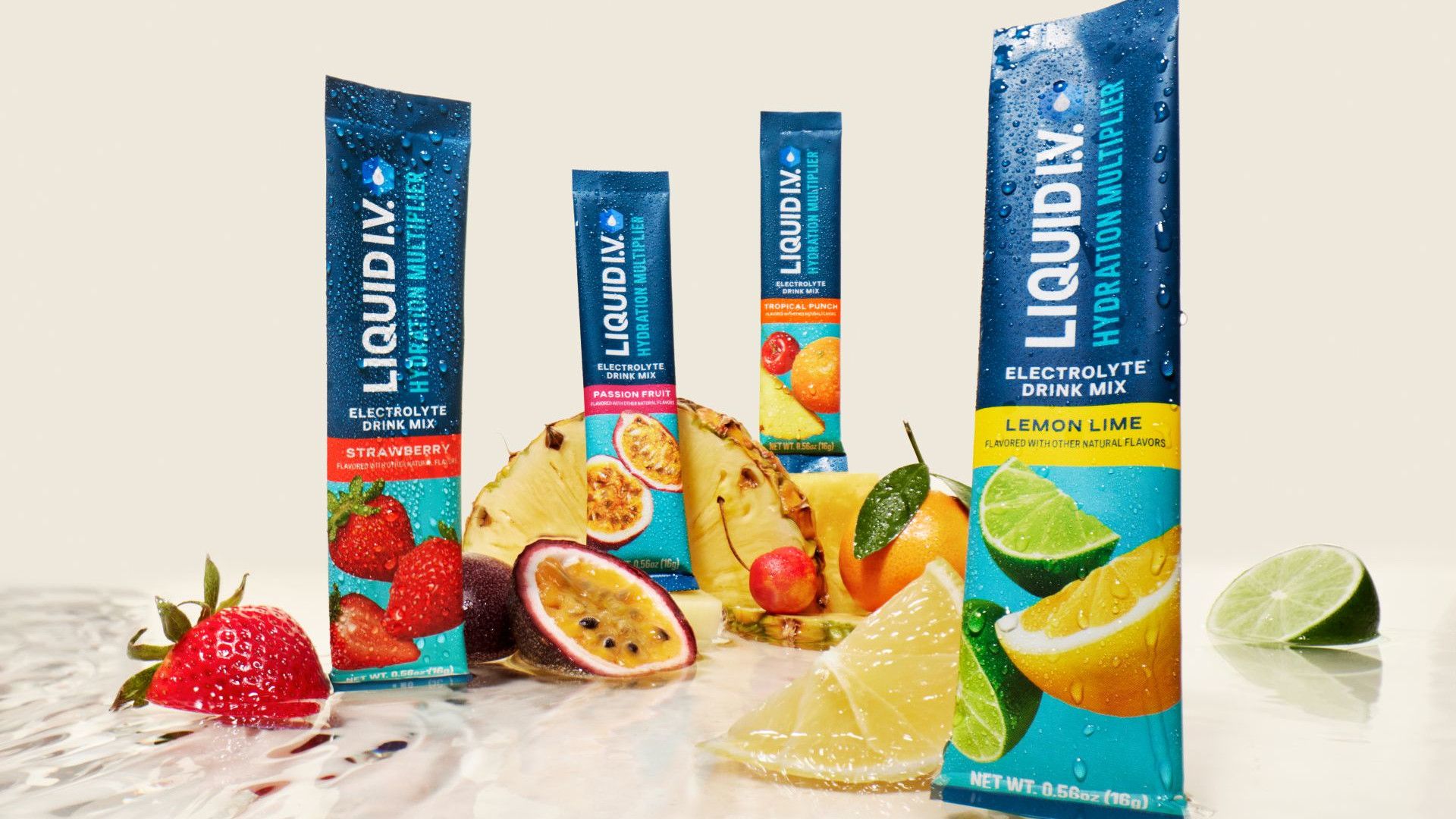 Assorted Liquid I.V. hydration multiplier sticks in different flavours, including strawberry, tropical punch, and lemon lime, displayed upright among fresh fruits such as strawberries, passion fruit, pineapple, citrus slices, and limes on a reflective surface.