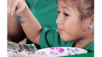 A female child eats while holding a spoon. In front of her is rice and a soup bowl