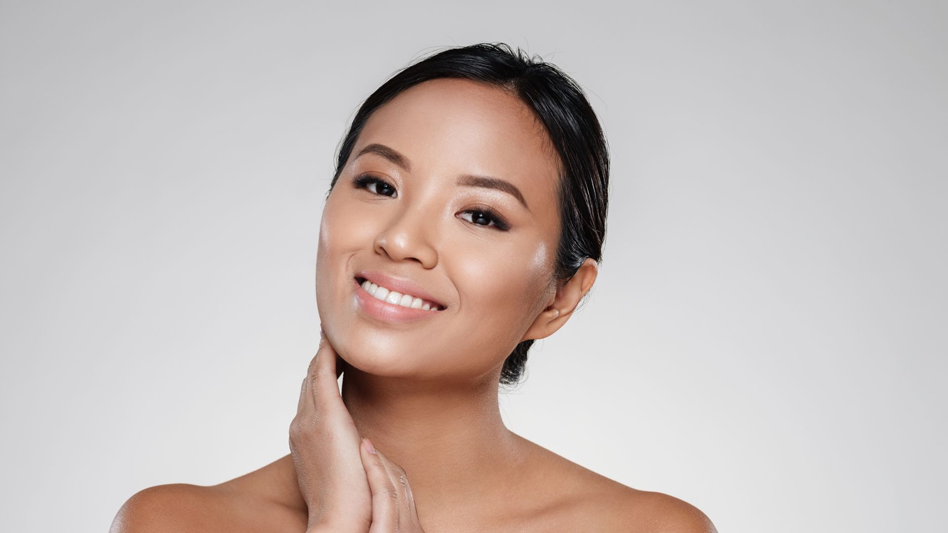 A Filipina woman is smiling at the camera while gently placing her hands near her cheek and neck.