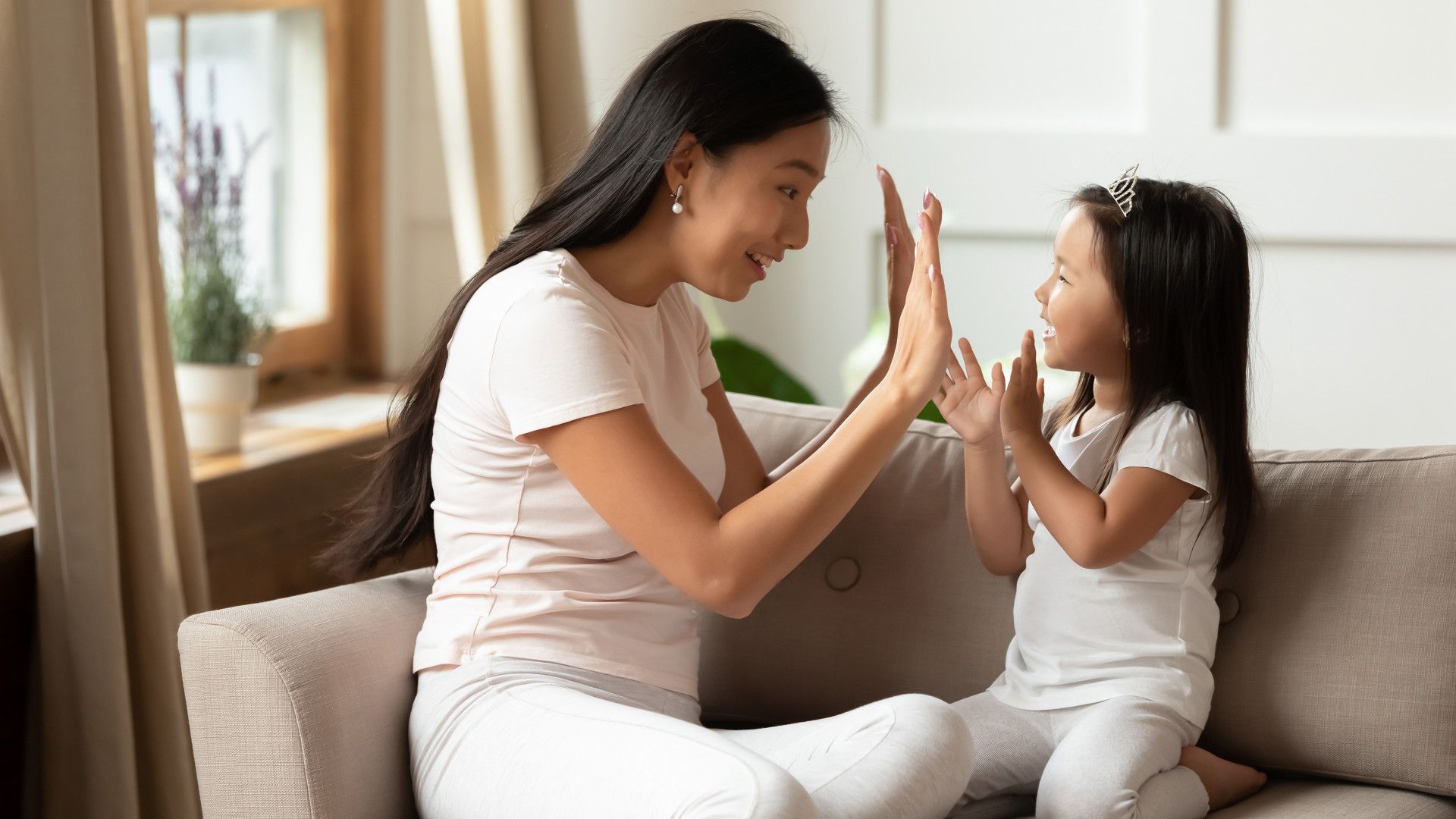 A woman and young girl sitting on a couch indoors, giving each other a high five in a bright living room.
