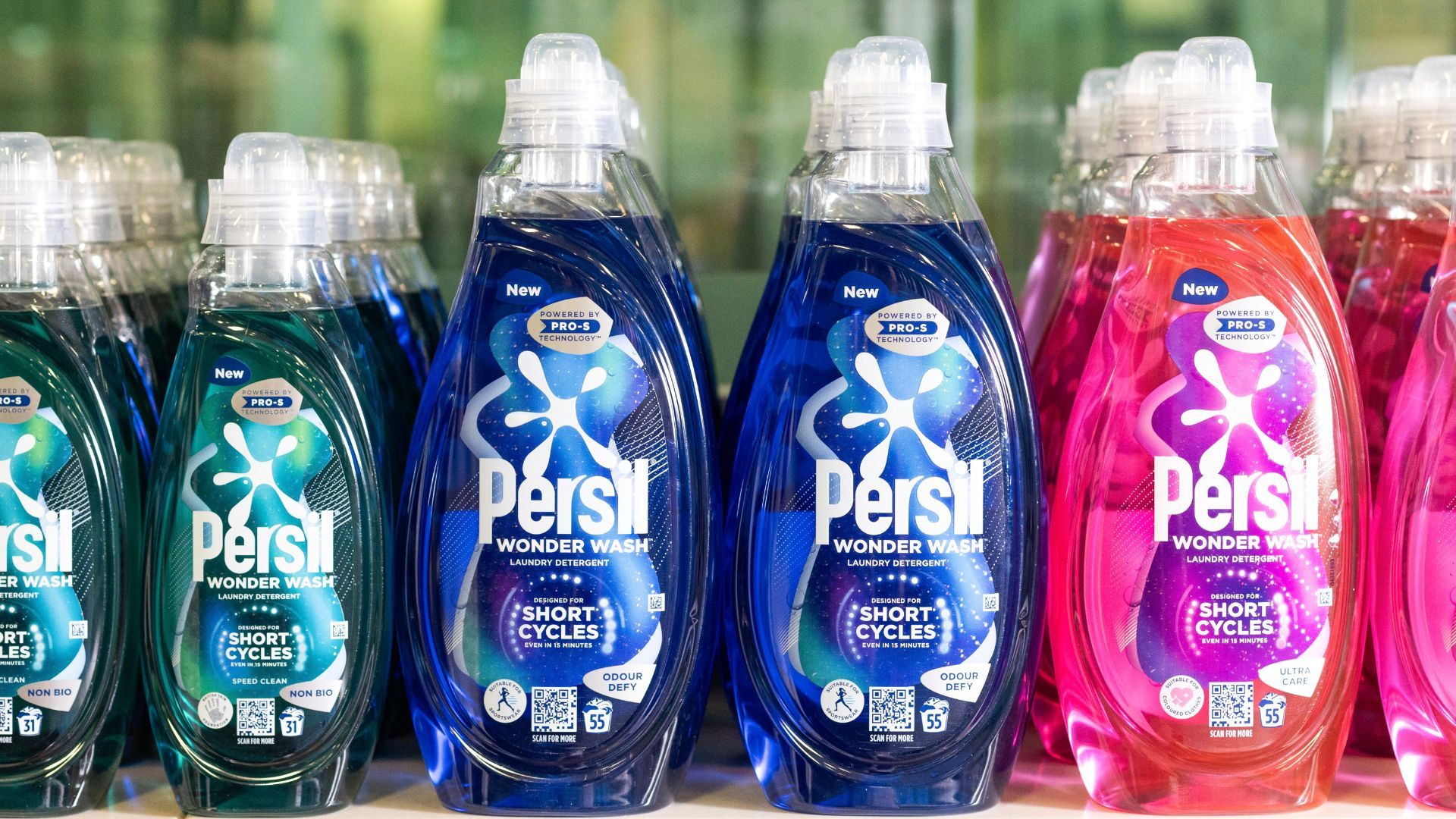 Persil Wonder Wash plastic laundry detergent bottles lined up in a row. There are three colours of detergent – green, blue and pink. The packaging labels explain that the product is designed for short cycles.